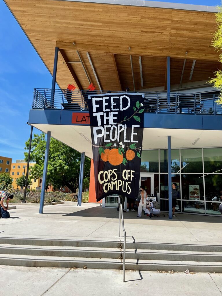 A black painted banner hangs from the balcony of Latitude dining commons, nearly reaching the ground below. It states 'FEED THE PEOPLE' and 'COPS OFF CAMPUS' in white paint. Between the two chunks of text is a branch with oranges and small white flowers