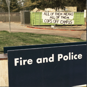 banner reading "all of them means all of them/cops off campus" on dumpster behind "Fire and Police" sign at UCD station