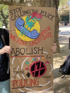 Painted cardboard sign reading "Stop calling the police. We keep each other safe. Abolish all forms of policing."