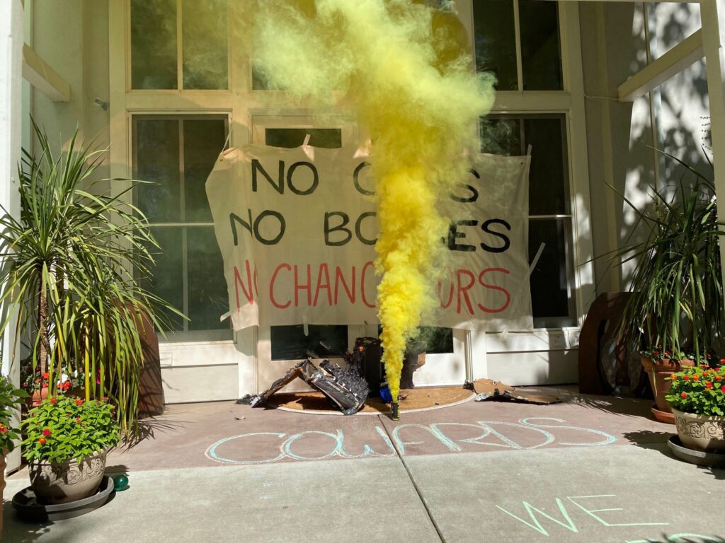 Banner outside Chancellor Gary May's residence reading "No cops no bosses no chancellors"; colorful yellow flare