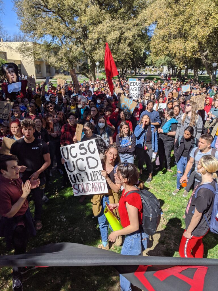 Crowd with protester at center holding sign reading "UCSC UCD UC-wide solidarity"