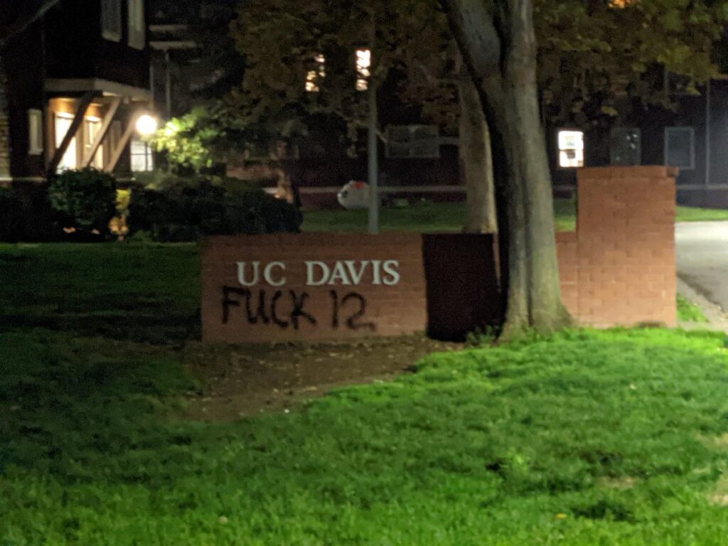 an image of a sign on the UC Davis campus. A brick wall at the entrance to campus has a sign that reads "UC Davis." Beneath this sign is spray painted the words "Fuck 12"