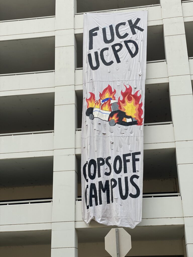 banner drop from garage next to police station: "fuck UCPD/Cops Off Campus" with image of burning police car