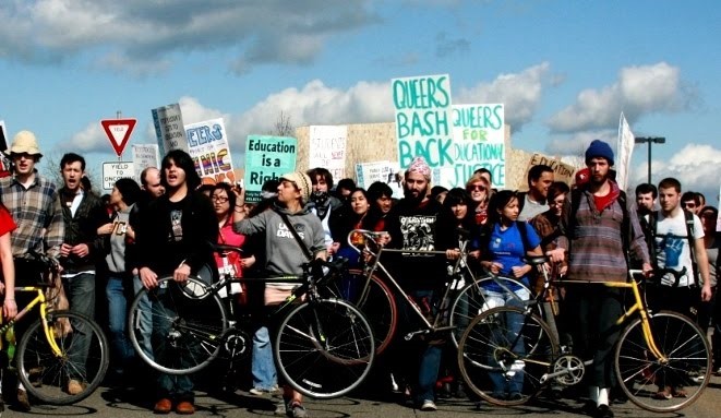 a photo of a crowd of protesters blocking a street near Davis California. They have a wall of bicycles that they are moving forward with them as they march, and carry signs reading "queers bash back" and other slogans
