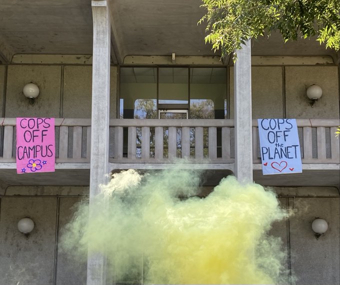 Pink banner reading "Cops off Campus"; white banner reading "Cops off the Planet" dropped from Olson Hall balcony; colorful yellow flare